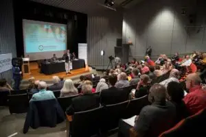 Theater room filled iwth people and table on stage with man standing up and speaking.