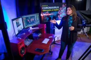 Woman gesturing to computer desk with speaker and 2 monitors set up with information on Covid 19 pulled up.