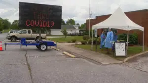 Volunteers in protective gear standing under pop up tent next to LED road sign that says "Drive Thru Covid19"