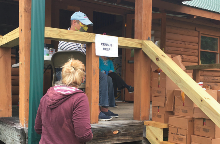 Woman approaching man on porch of log cabin with a sign that says "Census Help" and boxes stacked on the stairs