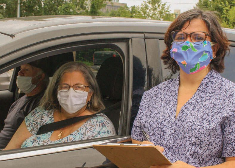 Female volunteer with facemask and clip board standing next to a car with window down and two people inside.