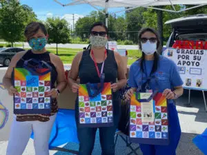 3 female volunteers from Hola Carolina holding up colorful reusable grocery bags while wearing masks at a pop up tent event.