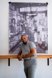 J Hackett leaning agains wooden railing below large picture of historic black-owned Asheville business.