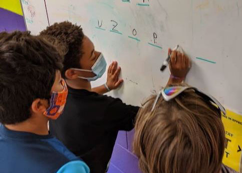 3 elementary students wearing facemasks with one writing letters on a white board.