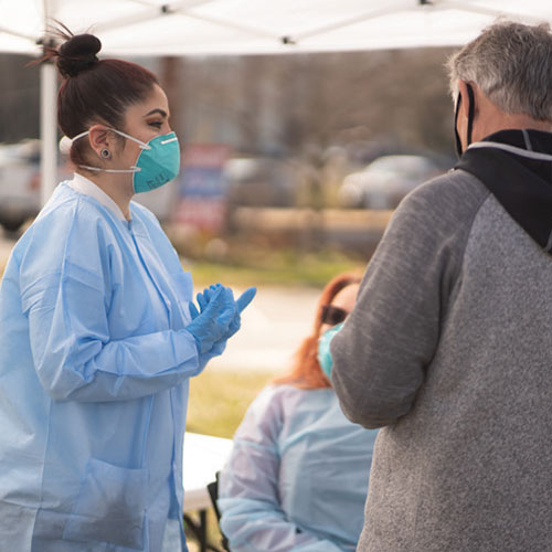 A woman wearing a face mask, gloves, and disposable lab coat talking with others in a pop-up tent outside.