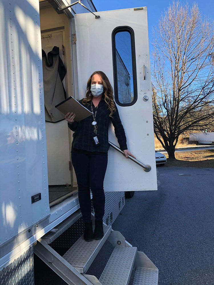 Woman wearing mask and holding writing pad stands on steps leading up to a mobile clinic.