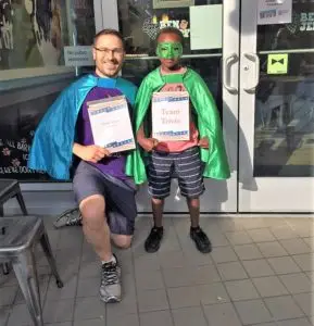 Man in blue cape standing with kid in green cape and eye mask holding up signs.