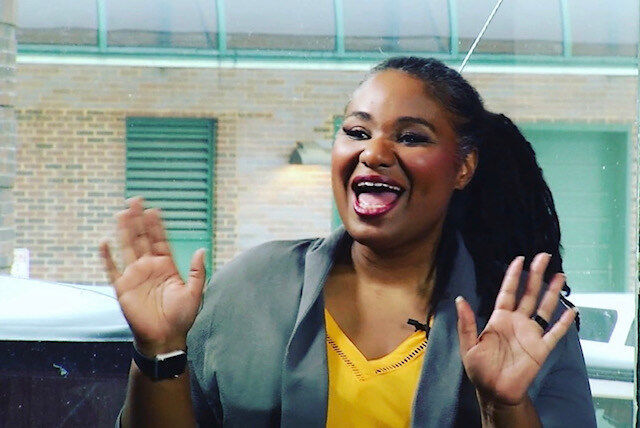 Woman in yellow blouse and grey blazer smiling with both hands up