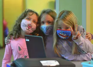 3 girls wearing fun facemasks smiling and front of school computer, one giving the peace sign.