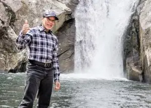 Older man with baseball cap standing in front of a North Carolina waterfall with a thumbs up.