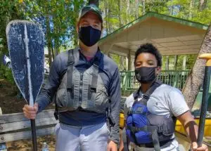 Man and young boy wearing masks while standing in fishing jackets and holding paddles.