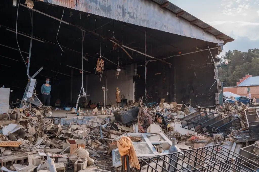 A large damaged warehouse filled with debris and rubble with two people are standing inside observing the scene
