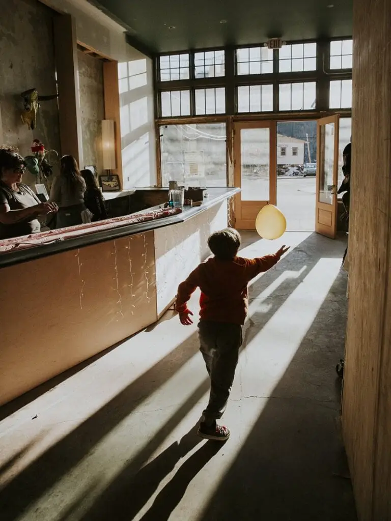 A child runs towards an open sunlit door, reaching for a floating yellow balloon as warm light casts long shadows inside the room