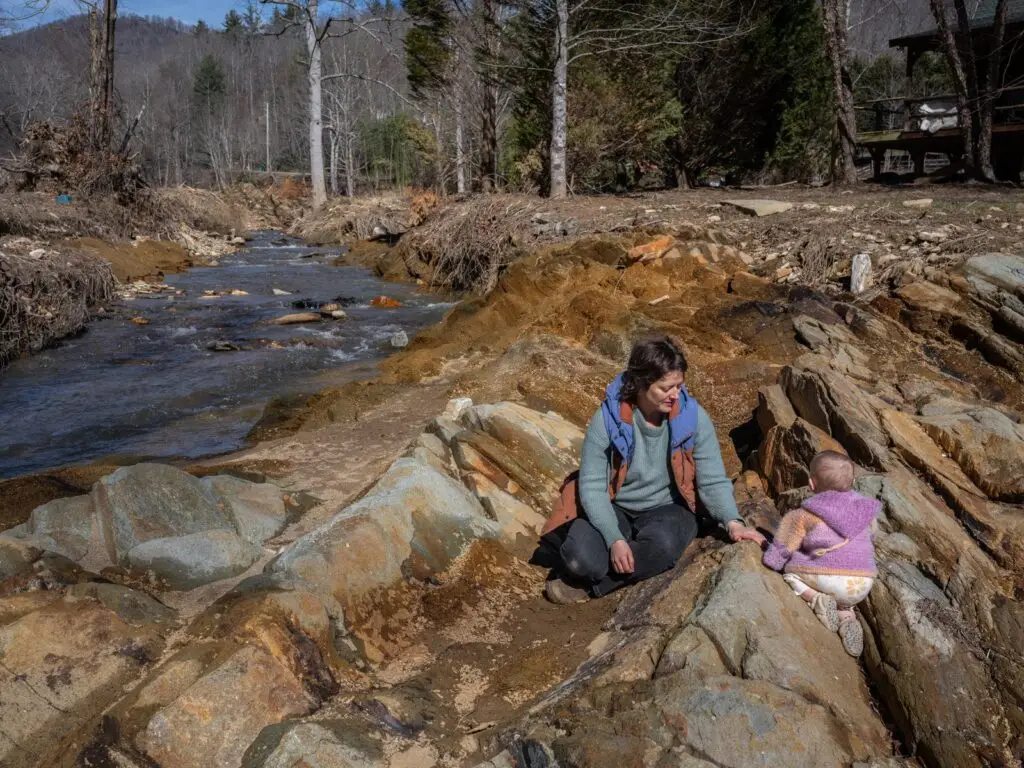 woman sits on rocky terrain beside a small child near a flowing creek with debris from hurricane helene
