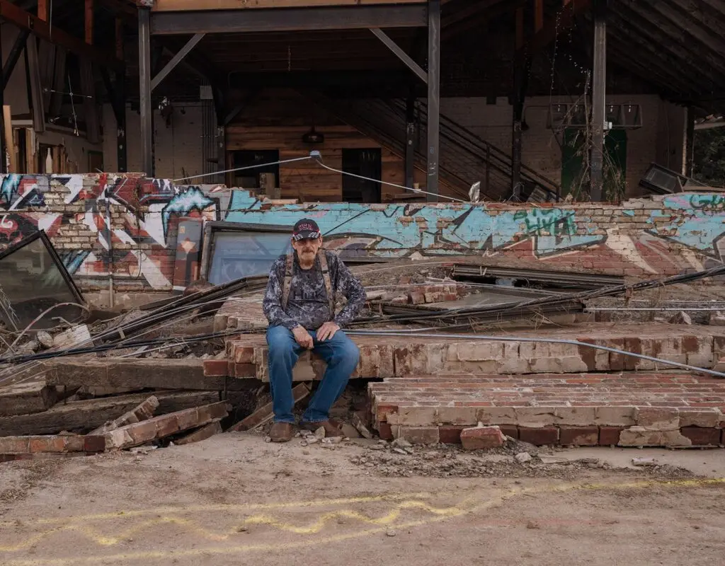 A man sits on rubble in front of a graffiti-covered building that was destroyed during Hurricane Helene