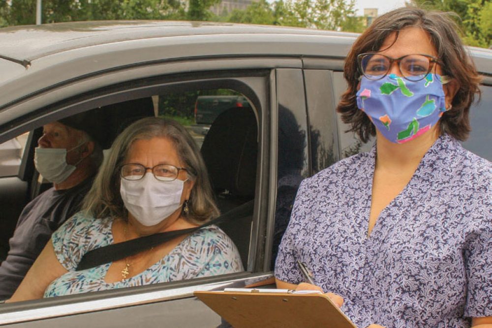 Female volunteer with facemask and clip board standing next to a car with window down and two people inside.