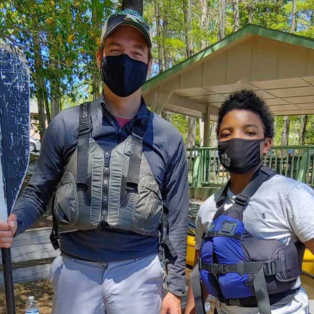 Man and young boy wearing masks while standing in fishing jackets and holding paddles.
