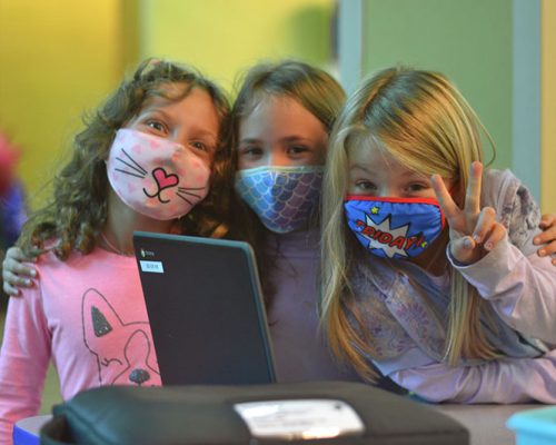 3 girls wearing fun facemasks smiling and front of school computer, one giving the peace sign.