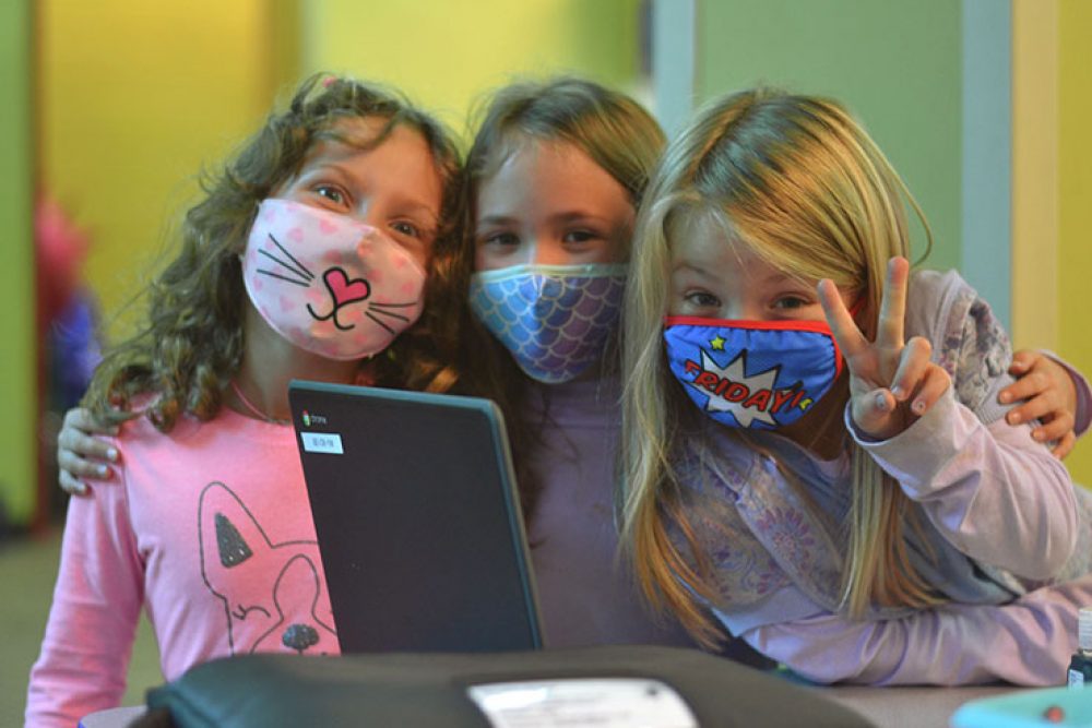 3 girls wearing fun facemasks smiling and front of school computer, one giving the peace sign.