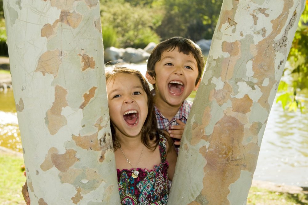 two children smiling from behind a tree