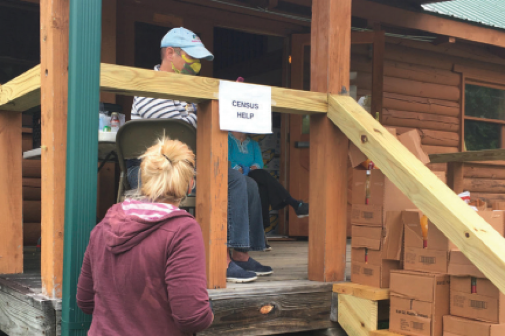 Woman approaching man on porch of log cabin with a sign that says "Census Help" and boxes stacked on the stairs