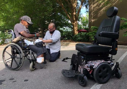 Man helping amputee in old wheelchair get ready for a new electric wheelchair.