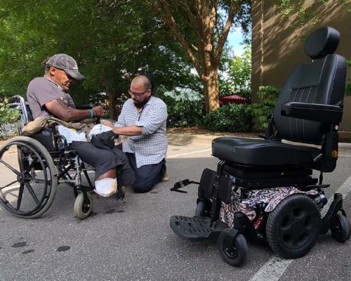 Man helping amputee in old wheelchair get ready for a new electric wheelchair.