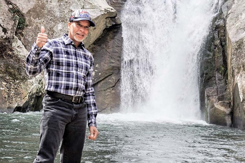 Older man with baseball cap standing in front of a North Carolina waterfall with a thumbs up.